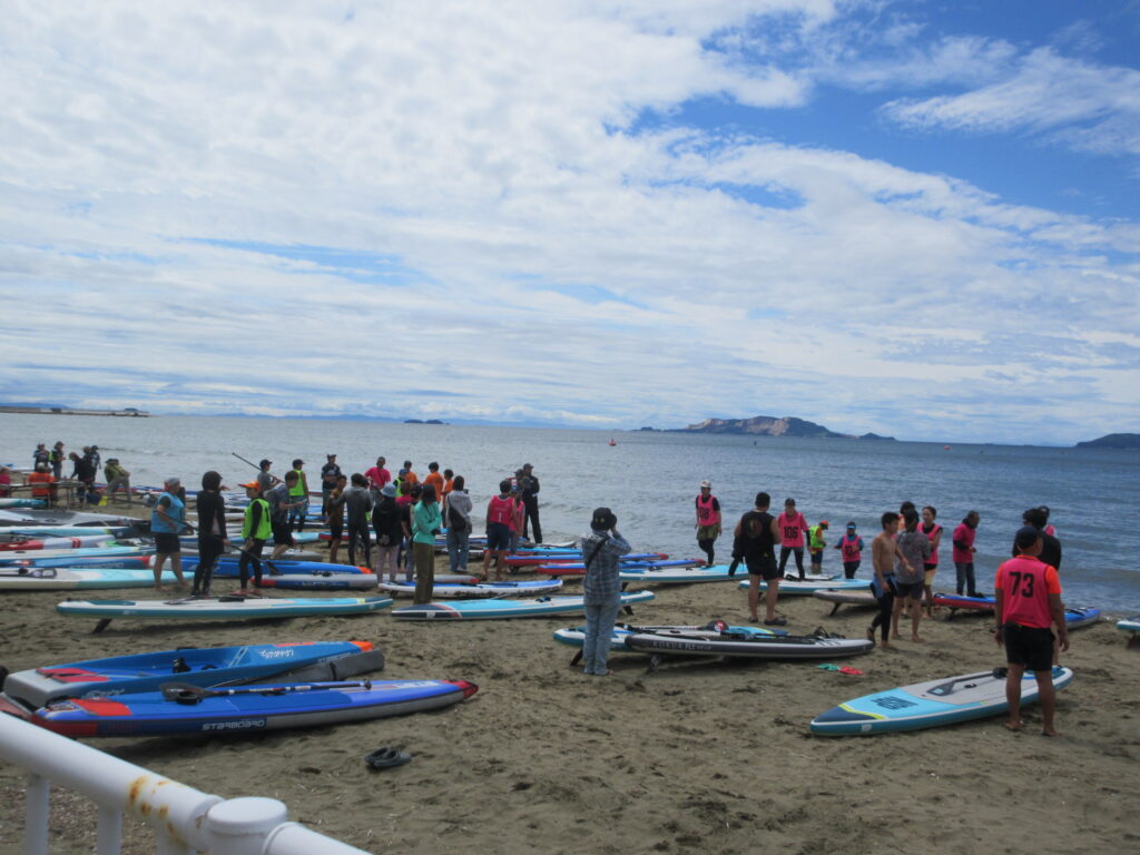 Crowded beach with many people near colorful stand-up paddleboards along the shore under a cloudy sky and calm water.