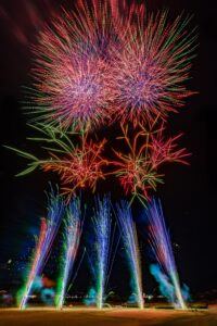 0019 1 - 【公式】兵庫県たつの市の観光サイト - 【公式】兵庫県たつの市の観光サイト Colorful fireworks explode in the night sky above a field, green and blue trails rising from ground-level fountains.