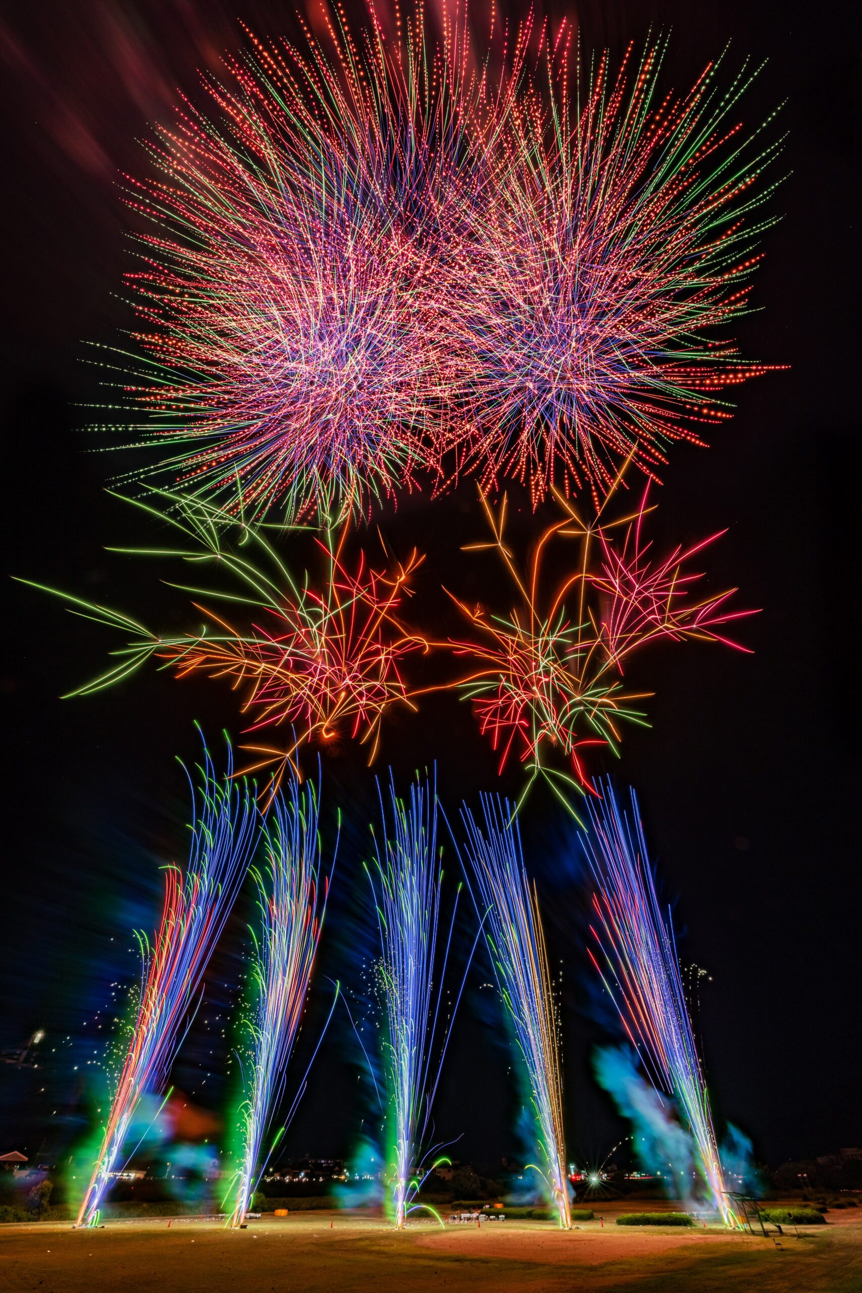 Colorful fireworks explode in the night sky above a field, green and blue trails rising from ground-level fountains.