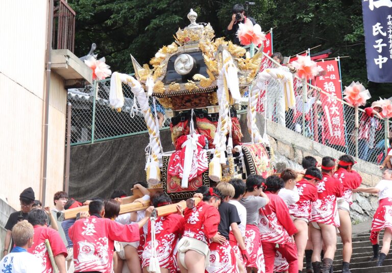 Crowd in red festival coats carry a richly decorated portable shrine (mikoshi) with gold details up stone steps during a Japanese matsuri, banners in the background.
