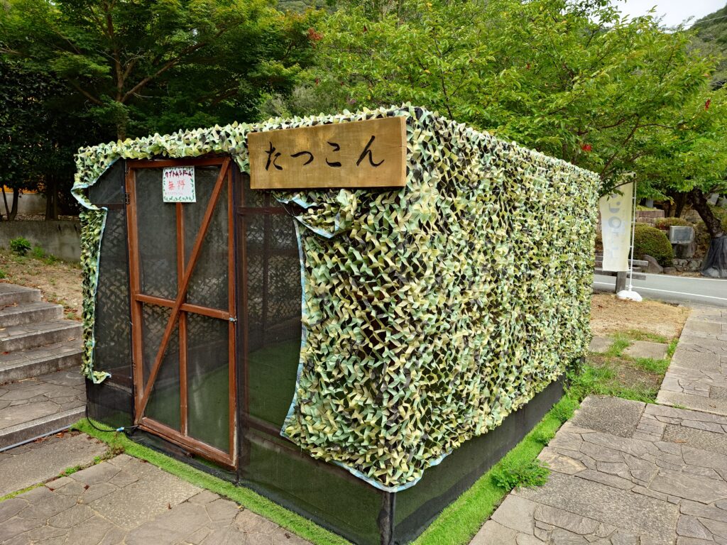 Wooden-framed shed wrapped in green camouflage netting in a park, with a wooden sign displaying the Japanese text たっこん.