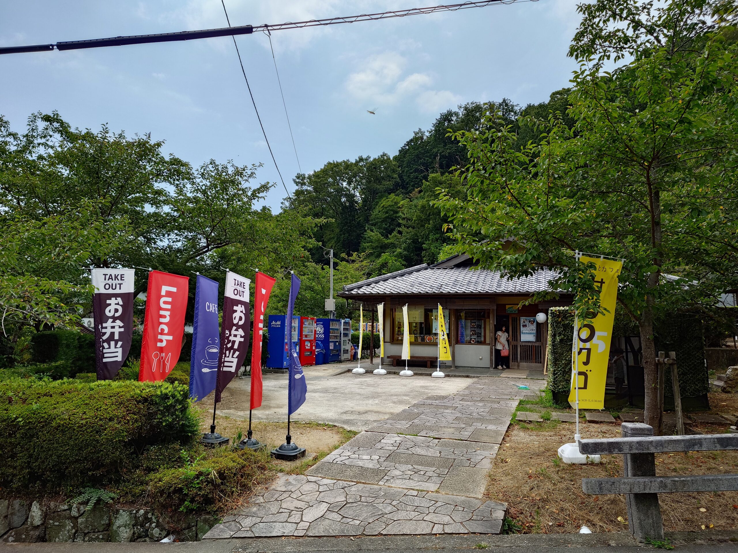 Row of tall vertical banners advertising takeout and lunch in a courtyard, with a small building and vending machines in the background among green trees.