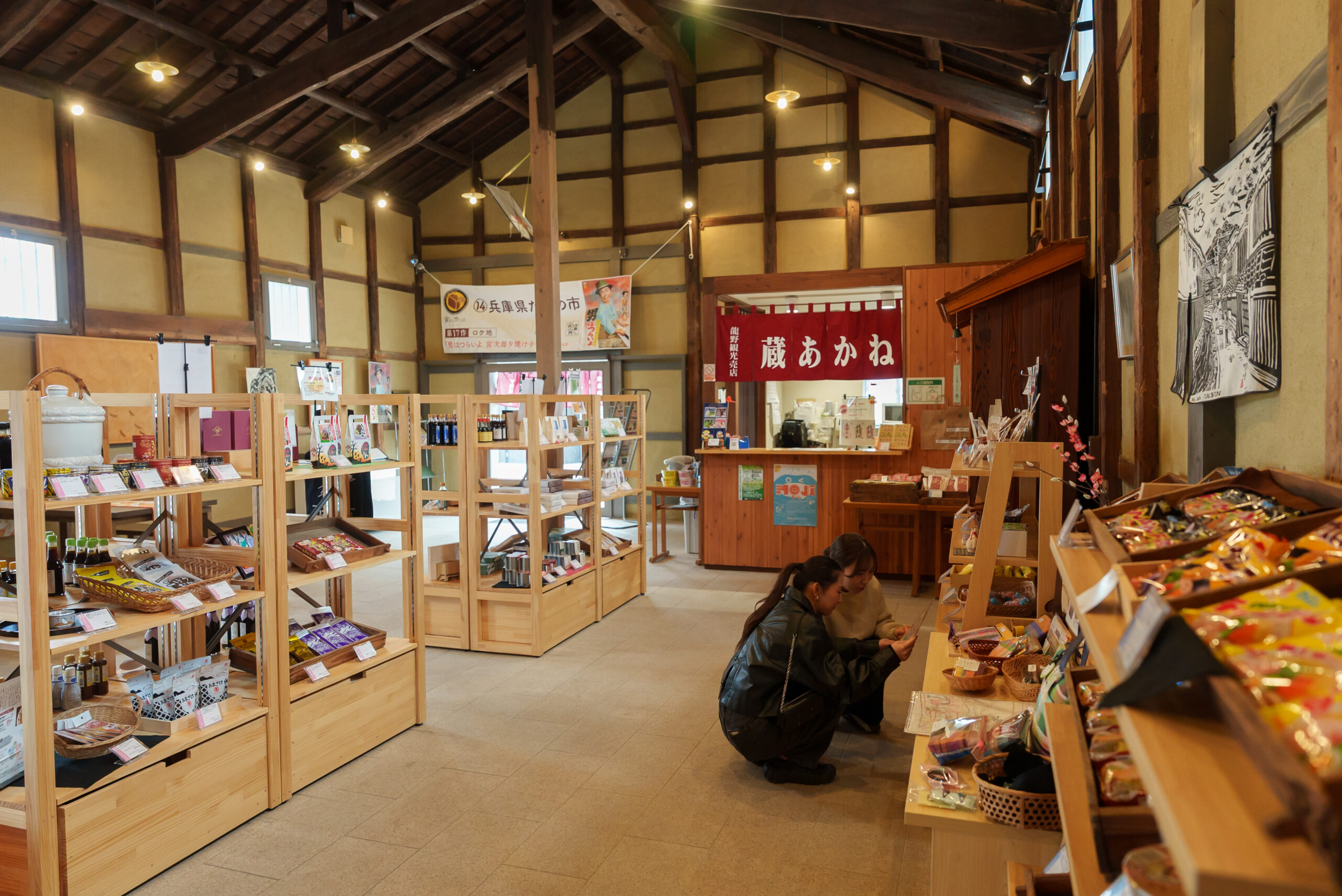 Interior of a rustic gift shop with wooden beams, shelves of products, and warm hanging lights; two people crouching and examining items on the floor in front of shelves.