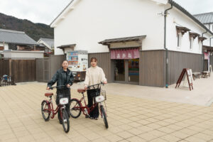 Two women standing with red bicycles in a plaza in front of a white building and vending machine.