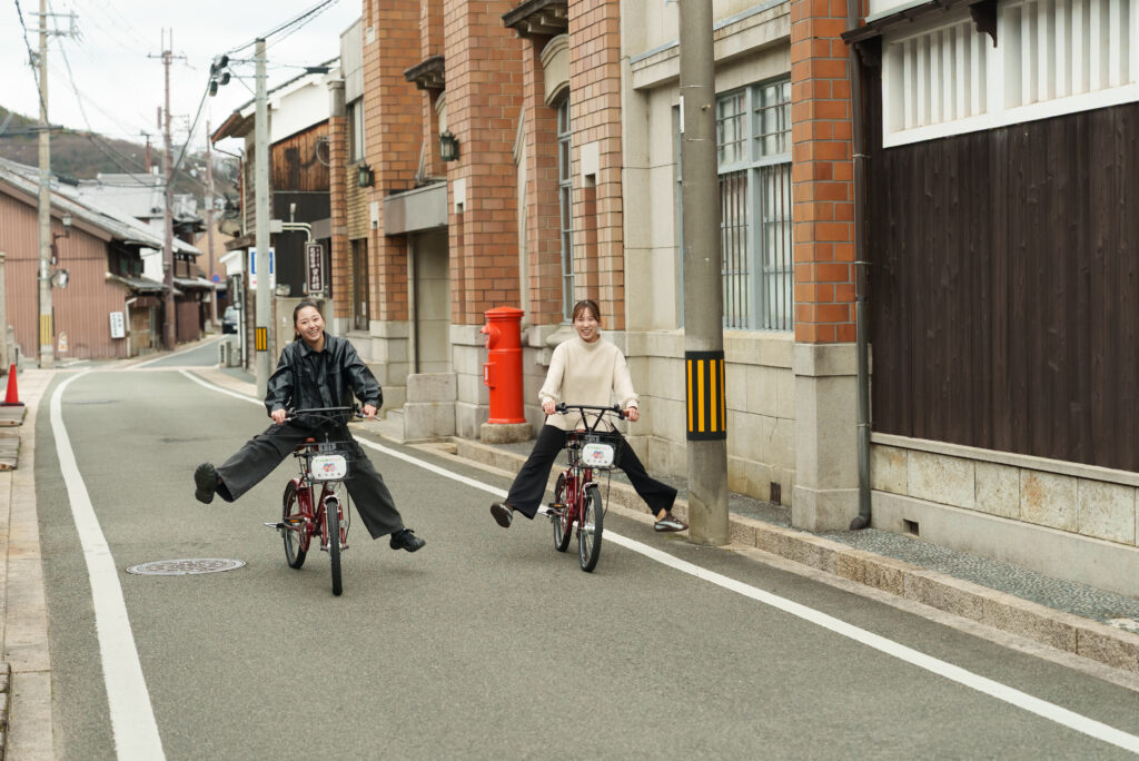 Two women ride bicycles down a narrow urban street; one is performing a wheelie and smiling.