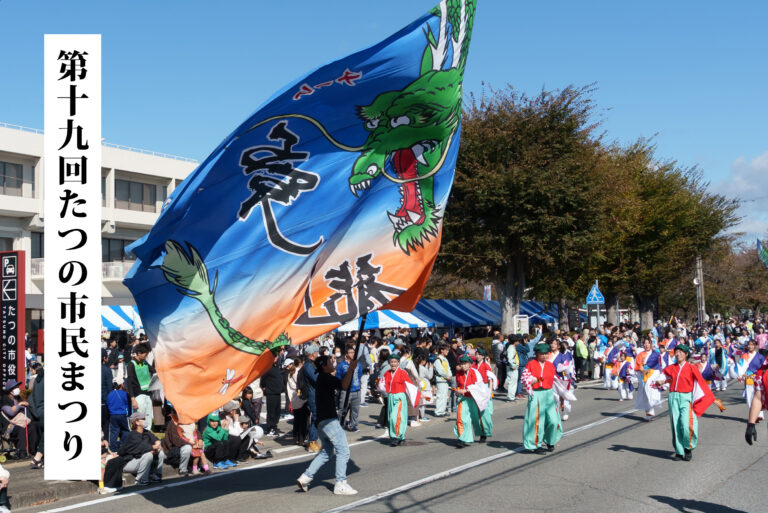 Large blue-orange dragon flag waves over a street parade with costumed performers and a cheering crowd; vertical Japanese banner on the left reads city event.